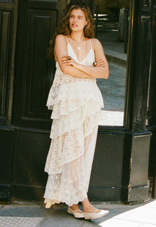 a-woman-stands-with-arms-crossed-wearing-a-white-lace-and-mesh-tiered-maxi-dress-with-spaghetti-straps-and-white-pointed-flats-posed-in-front-of-a-dark-wooden-doorway