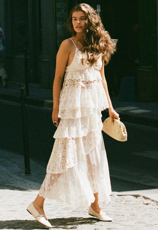 a-woman-walking-outdoors-in-a-white-lace-and-tulle-maxi-dress-with-a-tiered-ruffle-design-holding-a-small-beige-handbag-and-wearing-white-pointed-flats