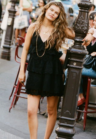 a-woman-wearing-a-black-tiered-ruffle-mini-dress-and-gold-necklaces-stands-on-a-city-street-next-to-a-dark-metal-post
