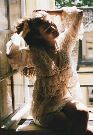 a-woman-wearing-a-sheer-white-lace-lingerie-set-with-ruffled-sleeves-and-a-matching-sheer-cover-up-sitting-by-a-sunlit-window