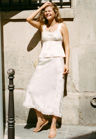 a-woman-wearing-a-white-sleeveless-lace-trimmed-top-and-matching-midi-skirt-stands-outdoors-against-a-sunlit-stone-wall-holding-a-small-beige-handbag-and-wearing-strappy-flat-sandals