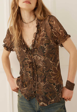 A woman wearing a brown and beige paisley patterned sheer blouse with ruffle trim, paired with blue jeans.