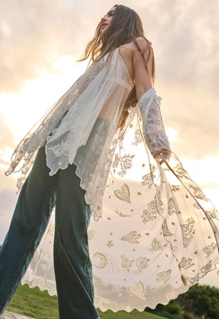 A woman outdoors wearing a sheer white lace long-sleeve cardigan with gold butterfly and moon motifs over dark teal wide-leg jeans